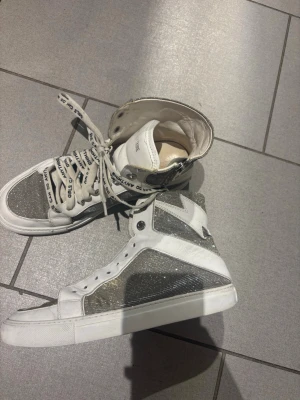 Glittriga vita high top sneakers - Säljer ett par vita high top sneakers från Zadig & Voltaire med silverglittriga paneler och en blixtdetalj på sidan. Skorna har vita snören med svart text och en cool, ungdomlig vibe. Perfekta för dig som vill sticka ut med din stil.