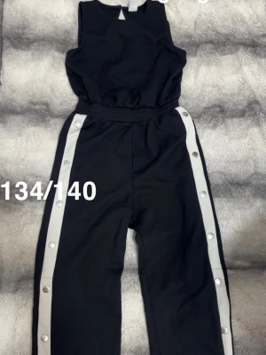 Svart jumpsuit med vita revärer - Snygg svart ärmlös jumpsuit med breda vita revärer längs benen och tryckknappar. Resår i midjan för skön passform och rund halsringning. Perfekt för dig som vill ha en sportig och trendig look.🌸 Använd två gånger. Storlek 134/140