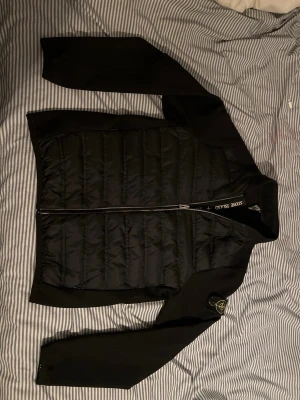 Svart dunväst från Stone Island - Snygg svart dunväst från Stone Island med quiltad front och dragkedja. Västen har klassisk Stone Island-logga på ärmen och detaljer i svart tyg. Perfekt för lager-på-lager och streetwear-stil. Den är i L med passar storlek M