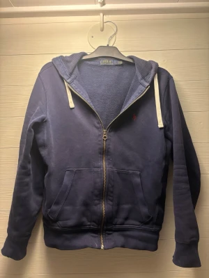 Marinblå hoodie från Polo Ralph Lauren - Marinblå hoodie med dragkedja från Polo Ralph Lauren. Klassisk design med huva och vita snören, två fickor framtill och broderad logga på bröstet. Tillverkad i mjuk bomullsblandning som är skön att ha på sig.