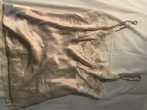 Champagnefärgat linne med spets - Sidenliknande llinne i champagnefärg med tunna axelband och vacker spetsdetalj vid bysten. Spetsen har blommönster och små pärlor som ger en lyxig känsla. Perfekt för dig som vill ha något elegant.