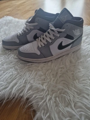 Nike Air Jordan 1 Mid grå/vit - Snygga Nike Air Jordan 1 Mid sneakers i grått och vitt med svart swoosh. Klassisk high-top design med perforerad tåbox, vita skosnören och ikoniska Air Jordan-loggan på sidan. Ovandelen är i läder och textil för en clean och sportig look.