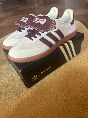 Adidas Wales Bonner Samba sneakers - Unika Adidas Wales Bonner Samba sneakers i vitt och vinrött med klassiska tre ränder på sidan. Skorna har en gummisula, detaljer i läder och textil samt en oversized plös med Wales Bonner-logga. Snygg retrostil och coola kontrastdetaljer.