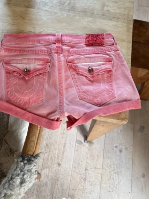 Rosa jeansshorts från True Religion - Säljer ett par snygga rosa jeansshorts från True Religion med uppvikta benslut och klassiska bakfickor med lock och knapp. Shortsen har låg midja och coola sömdetaljer. Perfekta för varma dagar och en avslappnad stil.