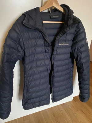 Svart dunjacka Peak Performance - Snygg svart dunjacka från Peak Performance med huva och dragkedja framtill. Jackan har diskret logga på bröstet och ärmen, samt två sidofickor med dragkedja. Perfekt för kalla vinterdagar tack vare det värmande dunmaterialet.