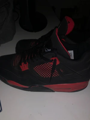 Svarta och röda Air Jordan 4 sneakers - Snygga Air Jordan 4 sneakers i svart och rött med klassisk Jumpman-logga på hälen. Skorna har meshpaneler på sidorna, röda detaljer och svart snörning. Ovandelen är i mocka och textil, och innersulan är röd med Air Jordan-etikett. Med insulorna som är lite skadade men känns helt perfekta 