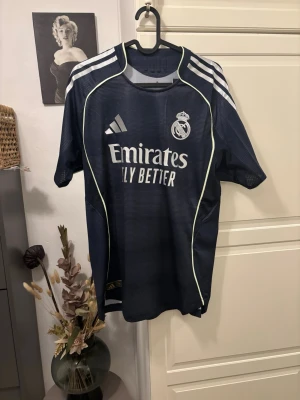 Real Madrid matchtröja mörkblå (authentic) - Snygg mörkblå Real Madrid matchtröja från Adidas med vita detaljer och klubbmärke på bröstet. Tröjan har korta ärmar, rund hals och klassiska Adidas-ränder på axlarna. Materialet är lätt och andas, perfekt för fotbollsträning eller match.