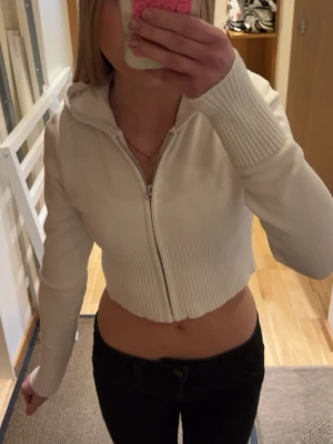 Vit croppad hoodie med dragkedja - Säljer en vit croppad hoodie med dragkedja framtill och ribbade muddar. Hoodien har en huva och är gjord i ett mjukt, stickat material som känns skönt mot huden. Perfekt till jeans eller kjol för en avslappnad och trendig look. Barnstorlek men skulle säga att den passar xxs/xs.