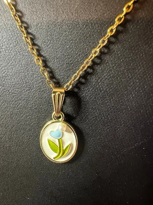 Guldhalsband med blomma och pärldetalj - Unikt halsband i guldfärgad metall med en oval berlock som har ett blommotiv i blått och grönt. Berlocken pryds av en liten pärldetalj upptill och har en emaljerad yta som ger en färgglad och lekfull känsla. Perfekt för dig som gillar söta och personliga smycken.