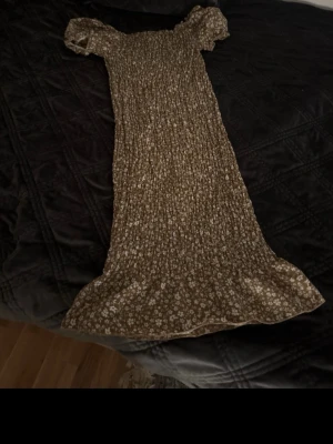 Brun blommig långklänning - Supersöt brun långklänning med små vita blommor och puffärmar. Klänningen har en smockad överdel och är tillverkad i ett mjukt, lätt tyg som känns perfekt för varmare dagar. Passformen är normal och den har en romantisk vibe med sina fina detaljer.