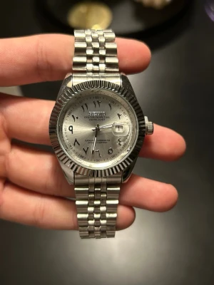 Seiko mod, custom made - Stilren Seiko Oyster Perpetual Datejust med silverfärgad urtavla och arabiska siffror. Klockan har en klassisk länk i rostfritt stål och datumfönster med förstoringsglas. Boetten har räfflad kant och hela klockan har en elegant, tidlös look. Custom made med urtavala/armband. Nh35 movement