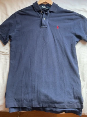 Marinblå pikétröja från Ralph Lauren - Klassisk marinblå pikétröja från Ralph Lauren med röd broderad logga på bröstet. Tröjan har korta ärmar, krage och två knappar framtill. Nypris 1300 ungefär. Knappt andvänd, därav inga defekter. Mitt pris: 350