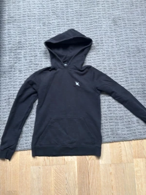 Axel Arigato Hoodie - Svart hoodie från Axel Arigato med liten vit logga på bröstet. Klassisk modell med huva och känguruficka framtill. Tillverkad i mjukt bomullsmaterial och har ribbade muddar vid ärmslut och nederkant. Perfekt för en avslappnad och stilren look.
