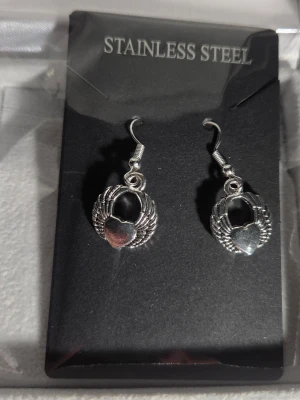 Silverörhängen med hjärta och vingar - Snygga örhängen i rostfritt stål med ett hjärta i mitten och detaljerade vingar runt om. De har en klassisk krok och en cool, silvrig finish som passar till många olika stilar. Perfekta för dig som gillar smycken med symboliska motiv.