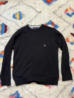 Svart Tommy Hilfiger tröja - Klassisk svart vintage Tommy Hilfiger tröja med U-ringning och broderad detalj på bröstet. Tröjan har ribbade muddar vid ärmslut och nederkant samt en diskret randig insida vid halsen. Perfekt för lager på lager och enkel att matcha. Storlek M men passar även som L