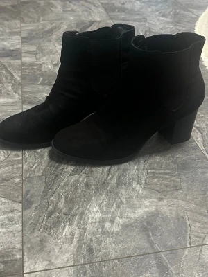 Svarta boots med klack - Snygga svarta stövletter i mockaimitation med rundad tå och bred klack. Skorna har en enkel och stilren design med resår på sidorna för enkel påtagning. Perfekta till jeans eller kjol för en trendig look. Klacken är 6-7 cm.