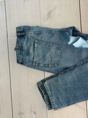 Blå raka jeans från Jack and Jones - Ett par klassiska blå jeans från Jack and Jones med rak passform och fem fickor. Jeansen har en ljus tvätt och subtila slitningar för en avslappnad look. Tillverkade i bomull med knappgylf och bälteshällor. Perfekta för en casual stil.