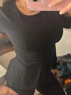 Svart peplum topp med kort ärm - Säljer en svart peplum topp med rund halsringning och korta ärmar. Toppen har en markerad midja som ger en snygg siluett och är tillverkad i ett mjukt, stretchigt material. Perfekt att styla med jeans eller kjol för en trendig look.