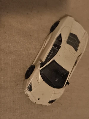 Vit leksaksbil sportmodell - Säljer en vit leksaksbil i sportbilsmodell med svarta detaljer och genomskinliga fönster. Bilen har en låg och aerodynamisk form, svarta hjul och detaljerade linjer på motorhuven och taket. Perfekt för dig som gillar bilar och samlar på modellbilar.