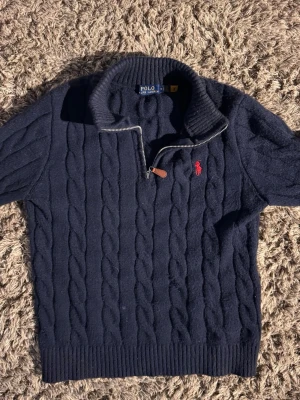 Mörkblå kabelstickad tröja Polo Ralph Lauren🙌🤩 - Mörkblå kabelstickad tröja från Polo Ralph Lauren med halv dragkedja och hög krage. Tröjan har ett rött broderat logomärke på bröstet och ribbstickade muddar. Perfekt för dig som gillar klassisk och stilren design.