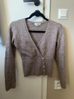 Beige omlottkofta från Gina Tricot - Säljer en beige stickad omlottkofta från Gina Tricot i alpackablandning, storlek xs. Koftan har långa ärmar, djup V-ringning och knäpps med fem knappar framtill. Perfekt för lager på lager och riktigt mysig att ha på sig. Knappt använd.