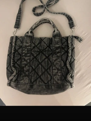 Grå quiltad handväska med axelrem - Snygg grå handväska i quiltad design med både handtag och avtagbar axelrem. Väskan har dragkedja upptill och ett rymligt svart innerfack. Materialet ser ut som tvättad denim vilket ger en cool och avslappnad look.