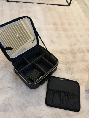 Svart sminkväska med spegel - Snygg svart sminkväska med hårt skal, inbyggd spegel och justerbara fack. Väskan har dragkedja, elastiska innerfickor och avtagbar insats för smart förvaring av smink eller accessoarer. Perfekt att ta med på resan eller ha hemma på sminkbordet.