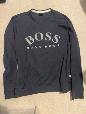 Mörkblå sweatshirt från Hugo Boss - Mörkblå sweatshirt med rund hals och stora vita BOSS-trycket framtill. Klassisk passform med ribbade muddar vid ärmslut och nederkant. Perfekt för en avslappnad och stilren look.