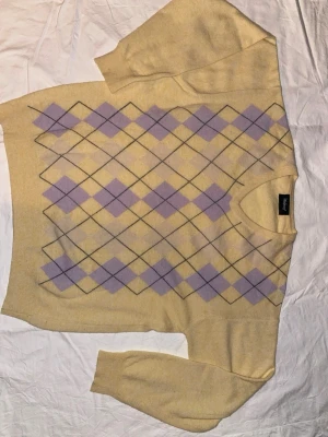 Gul stickad tröja med argylemönster - Snygg gul stickad tröja från Monki med klassiskt argylemönster i lila och svart på framsidan. Tröjan har rund halsringning och långa ärmar. Perfekt för dig som gillar retrovibbar och vill ha något mjukt och bekvämt i garderoben.