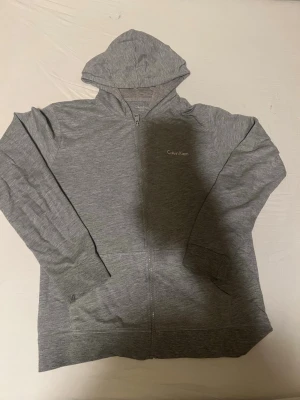 Grå zip hoodie från Calvin Klein - Snygg grå hoodie från Calvin Klein med dragkedja framtill och huva. Tillverkad i mjukt material och har två fickor på magen. Diskret Calvin Klein-logga broderad på bröstet. Perfekt för en chill och stilren look.
