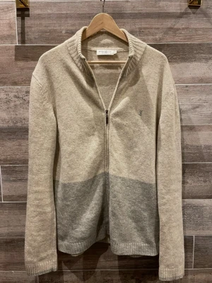 Yves Saint Laurent zip - Stilren ziptröja från Yves Saint Laurent i mjuk ullblandning. Tröjan är beige upptill och ljusgrå nedtill, har ribbad krage och muddar samt diskret YSL-logga broderad på bröstet. Perfekt för lager på lager och en clean look. Nypris 6000