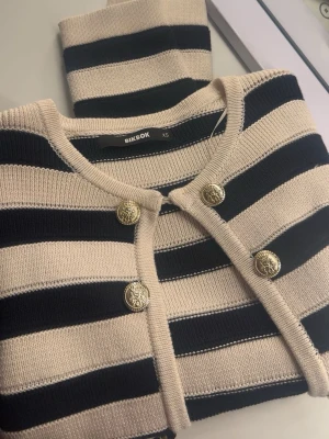 Randig stickad kofta från BikBok - Snygg stickad kofta från BikBok i beige och svart med breda ränder. Koftan har rund halsringning och fyra guldiga dekorativa knappar framtill. Perfekt för lager på lager och ger en trendig look. Helt ny och aldrig använd. Slutsåld. Köpt för 399