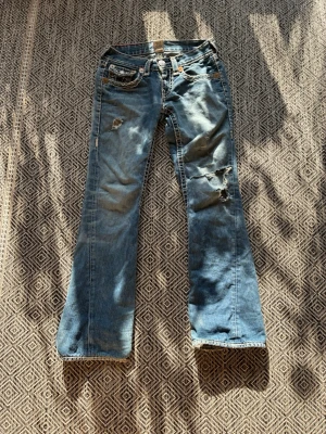 Blå slitna bootcut jeans True Religion - Säljer ett par blå bootcut jeans från True Religion med slitningar och hål på framsidan. Jeansen har klassiska kontrastsömmar, fem fickor och detaljerade bakfickor med knapp. Tillverkade i denim med en cool vintage look Lite trasiga i bak där den har tappat en knap..