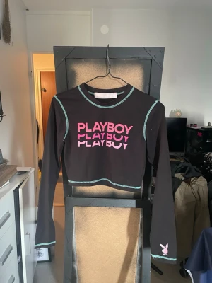 Svart croppad topp Playboy x Missguided - Snygg svart croppad långärmad topp från Playboy x Missguided. Toppen har turkosa kontrastsömmar, rosa PLAYBOY-tryck framtill och en liten kaninlogga vid ärmslutet. Perfekt för dig som vill sticka ut med en trendig streetstyle-look.