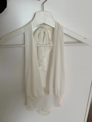 Vit djup urringad topp med knyt - Säljer en vit topp med djup urringning och knyt i nacken. Toppen har en mjuk, något transparent känsla och är ärmlös med breda axelband. Perfekt för dig som vill ha en stilren och trendig look. Materialet känns lätt och följsamt.