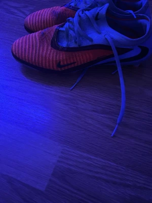 Nike orangea fotbollsskor med snörning - Säljer ett par snygga orangea fotbollsskor från Nike med svart swoosh och vita snören. Skorna har en låg profil och är designade för bra grepp på planen. Ovandelen är i syntetmaterial med textur för bättre bollkontroll. Perfekta för dig som vill sticka ut på fotbollsplanen.