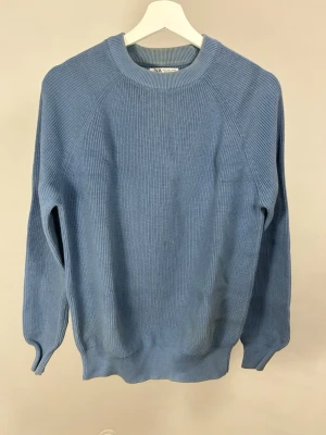 Blå stickad tröja från Zara | S - Snygg blå stickad tröja från Zara med rund halsringning och ribbade muddar vid ärmslut och nederkant. Missfärgad vid kragen och lätt defekt vid magen (se bilder).  Tröjan har en enkel, stilren design och är perfekt för dig som gillar en avslappnad men trendig look.