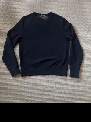 Burberry mörkblå sweatshirt med V-ringning - En stilren mörkblå Burberry sweatshirt med V-ringad hals och långa ärmar. Tröjan har ribbade muddar vid ärmslut och nederkant. Perfekt för en avslappnad och enkel look. Materialet ser ut att vara mjukt och bekvämt, troligen bomull eller bomullsblandning.