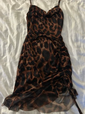 Leopardmönstrad kort klänning - Snygg kort klänning med leopardmönster i bruna och svarta toner. Klänningen har tunna axelband, figurnära passform och en dragsko med snörning på sidan för extra detalj.