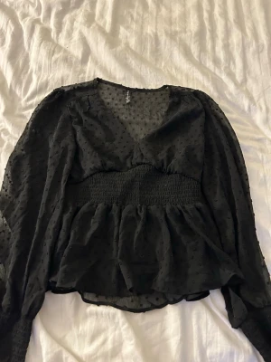 Svart transparent blus från Amisu - Snygg svart blus från Amisu med transparent tyg och diskreta prickar. Blusen har långärmade puffärmar, small midja och volang nedtill.