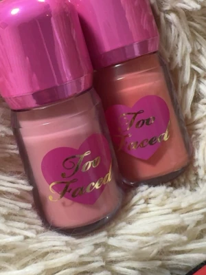 Too Faced Love Flush flytande rouge - Två flaskor flytande rouge från Too Faced i serien Love Flush. Färgerna är en mjuk rosa (Love Letter) och en varm persikoton (First Love). Flaskorna har rosa lock och hjärtformad etikett med guldtext. Perfekt för en fräsch och naturlig look.