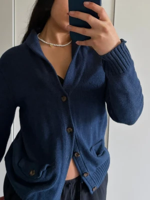 Mörkblå kofta med knappar - Mysig mörkblå kofta med stora bruna knappar och två fickor framtill. Koftan har ribbade muddar vid ärmslut och nederkant samt en avslappnad passform. Perfekt att slänga över en t-shirt för en chill look.