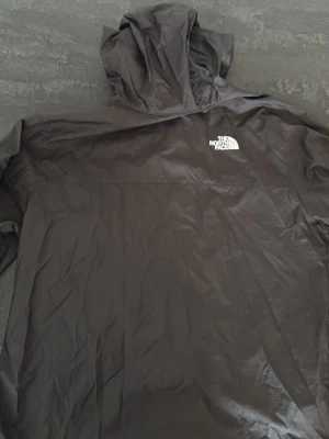 Svart vindjacka från The North Face - Snygg svart vindjacka från The North Face med huva och logga på bröstet. Tillverkad i lätt och tåligt material som skyddar mot vind. Perfekt för dig som vill ha en stilren och funktionell jacka till vardagsoutfiten.