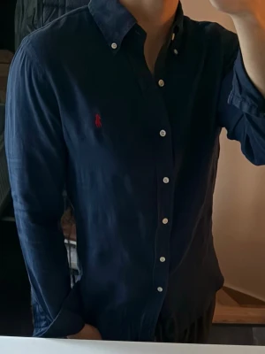 Mörkblå linneskjorta från Ralph Lauren - Klassisk mörkblå linneskjorta från Ralph Lauren med vit knappstängning och ikonisk röd logga broderad på bröstet. Skjortan har lång ärm, button-down krage och är tillverkad i 100% linne. Perfekt för en clean och stilren look. Lite urtvöttad annars bra skick