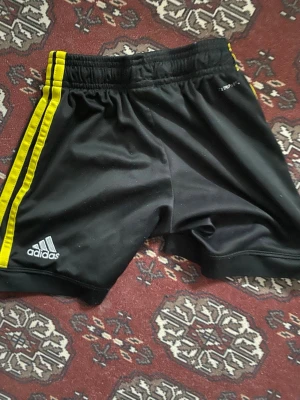 Svarta Adidas shorts med gula ränder - Säljer ett par svarta sportshorts från Adidas med gula ränder längs sidorna. Shortsen har tryckt Adidas-logga, Stadium-tryck och Hammarby IF-märke. Perfekta för träning eller match. Tillverkade i lätt och snabbtorkande material.