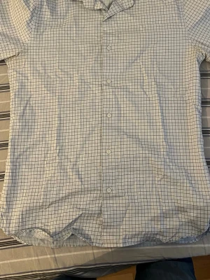 Vit rutig skjorta från Riley - Klassisk vit skjorta från Riley med tunna svarta rutor. Skjortan har knappar framtill, krage och lång ärm. Perfekt för en clean och stilren look. Tillverkad i bomull för en skön känsla hela dagen.