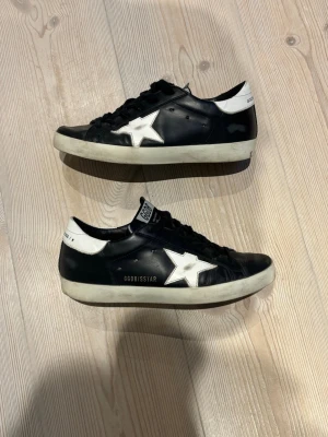Svarta Golden Goose stl 39 - Svarta sneakers från Golden Goose med vit stjärndetalj på sidan och vit hälkappa. Skorna har svart ovandel i läder, vita sulor och klassisk låg modell. Snygga kontraster och cool streetstil med GGDB/SSTAR-tryck på sidan.