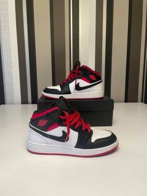 Nike Air Jordan 1 Mid  - Snygga Air Jordan 1 Mid sneakers från Nike i svart, vitt och rött. Klassisk design med röd yttersula, röda skosnören och ikoniska Swoosh-loggan på sidan. Tillverkade i läder med vadderad krage och perforerad tå för extra komfort. Skorna är som nya.