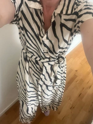 Zebramönstrad omlottklänning med knyt - Snygg långklänning med zebramönster i svart och vitt. Klänningen har omlottmodell, korta ärmar, krage och ett matchande knytband i midjan. Perfekt för dig som vill sticka ut med ett djuriskt print.