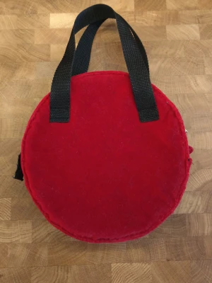 Rund röd handväska i tyg - Unik rund handväska i rött tyg med svarta, breda handtag och dragkedja upptill. Väskan har en enkel design utan synligt märke och är perfekt för dig som vill sticka ut med en annorlunda form. Materialet ser ut att vara mjukt tyg och dragkedjan är i metall. 20 diameter.